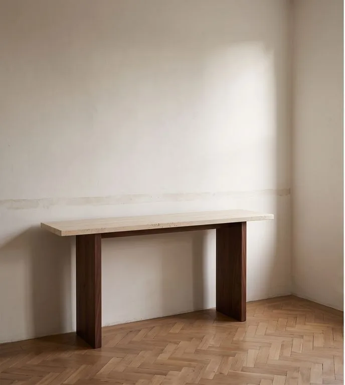 Long travertine console table on two solid walnut blade legs, against a plain wall on herringbone parquet.